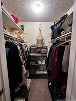 View of master closet showing hanging vintage clothing on both sides, shelving unit at back with books, accessories, and decorative lamp on top. Plush teddy bears are visible on upper shelf.