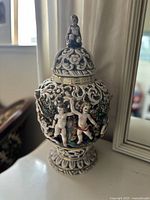 Front and side view showing the vase's sculpted cherubs and floral designs in relief, set on a pedestal base.