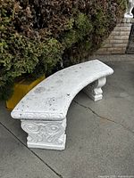 Vintage curved white garden bench with ornate scrollwork legs, placed on concrete surface near shrubbery.