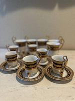 Seven vintage demitasse/espresso cups with matching saucers, showing gold and black decorative patterning with small footsteps and ornate handles on the cups.