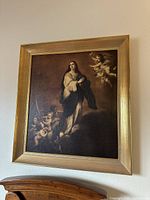 Full view of framed vintage religious artwork showing The Immaculate Conception with Virgin Mary and cherubs in gold-tone frame hanging on wall above wooden furniture.