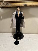 Full view of the Frank Sinatra porcelain doll standing on its black base with beige overcoat and black hat in front