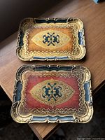 Pair of vintage Florentine trays, one with tan background and one with red background, showing ornate gold accents and blue borders.