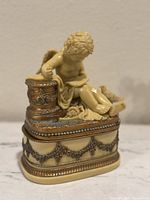 Front angle of vintage cream-colored resin cherub reading a book, seated on ornate decorative trinket box base with gold painted accents and bead detailing.