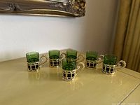 Set of six green glass shot glasses in gold-tone holders, placed on a surface showing their design and arrangement.