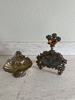 Side by side view of the gold filigree perfume bottle and soap dish on a white surface in front of a white wall.