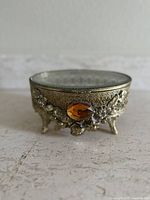 Side view of gold filigree trinket box showing amber gemstone and ornate floral metalwork with four feet.