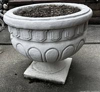 Front angled view of large vintage cement planter sitting on concrete ground showing woven pattern detail and pedestal base.