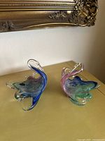 Pair of vintage Murano glass swans displayed on a yellow surface with a framed artwork in background, showing multicolored glass with blue, green, pink and clear accents.