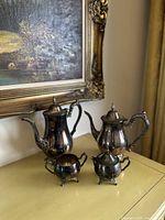 Full view of the vintage silverplate coffee and tea set with four pieces on a yellow surface beside a framed painting.