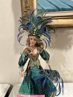 Front and side view of the doll wearing detailed green and gold costume with peacock feather headpiece and fan.