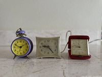 Set of three vintage alarm clocks displayed on a flat surface: a round purple metal bell alarm clock, a cream square clock, and a red foldable case travel clock.