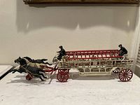 Full side view showing the vintage cast iron horse-drawn ladder fire truck with two black horses, red ladder and wheels, cream body, and two firefighter figurines.