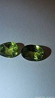Two natural apple green peridot gemstones laid side by side on a white surface under direct light, showing their oval and pear cut shapes and vivid green color.