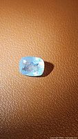 Photo showing the natural blue aquamarine gemstone on a textured surface, highlighting the stone's color, cushion cut shape, and internal inclusions.