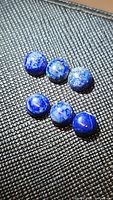 Six round deep blue cabochon lapis lazuli gemstones with white and gold speckles on black textured background