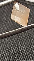 Close-up image of a natural peach moonstone gemstone with a fancy geometric cut held by tweezers against a black textured background. The image highlights the peach hue and faceted sides of the stone.