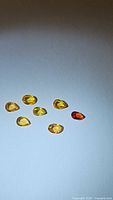 Seven natural sapphires in pear and oval cuts laid out on a white background, showing yellow, orange, and reddish hues.