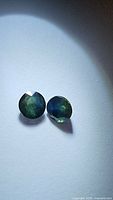 Pair of natural round cut blue-green sapphires shown on white background with lighting to highlight facets and color.