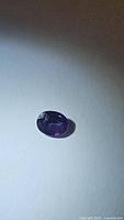 Photo of a purple spinel gemstone, oval cut, placed on a flat white surface showing its color and clarity under natural light.