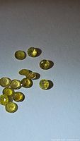 Top-down image showing 14 small natural yellow round cut tourmaline gemstones scattered on a white background under bright lighting.