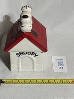 Front view of the ceramic Snoopy cookie jar with red roof and Snoopy figure lying on top of the doghouse, showing 'SNOOPY' text above door.