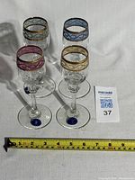 Set of four Cristalleria Fratelli Fumo crystal wine glasses with decorative gold gilt rims in pink, gold, and blue color accents, placed next to a measuring tape showing length approximately 8 inches.