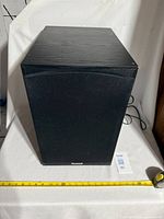 Front view of the Tannoy PS 110 subwoofer with black fabric grille and wood finish cabinet. Tape measure shows approximate size.