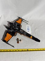Black and orange Lego Star Wars X-Wing Fighter with pilot minifigure and BB-series astromech droid placed next to it on fabric background with measuring tape.