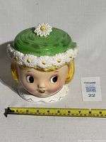 Front view of ceramic cookie jar shaped like a girl's head with a green lid decorated with white daisies and a daisy knob.