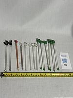 Full display of 13 vintage glass stir sticks showing different decorative tops including Playboy Bunny heads, olives, clear balls, and a pink twisted stick, placed next to a ruler for size reference.