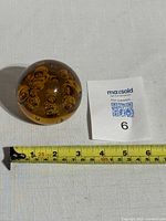 Top view of the amber glass sphere paperweight next to a MaxSold support card and measuring tape showing approximately 3.5 inches in diameter.
