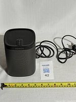 Front view of black SONOS PLAY:1 speaker with attached power cord and measuring tape showing size approximately 7 inches height.