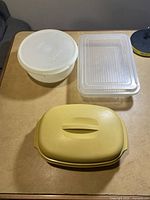Photo showing three assorted Tupperware containers positioned on a table: round white container with lid, rectangular clear container with lid, and yellow rectangular container with lid