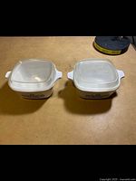 Two CorningWare square baking dishes with lids placed on a table, showing top view.
