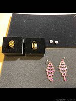 Overview of all jewellery items on black foam holders: two rings (one with yellow stone, one gold band ring), small round clear stone stud earrings, pink heart drop earrings.