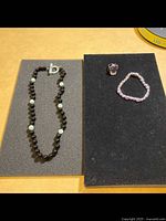 Photo shows a dark beaded necklace with white beads and a pink ring with a matching pink beaded bracelet on black display pads.
