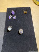 Full set of costume jewellery items on black felt background including purple stone earrings, white stone ring, blue stone filigree ring, and gold-tone clip-ons.
