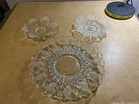 Photo showing three clear glass serving dishes on a table, including a large oval dish with leaf pattern and two smaller flower-shaped round dishes.