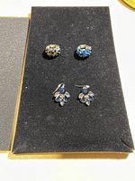 Two dome-style rings, one with clear stones and one with blue stones, along with earrings featuring blue and clear marquise-shaped stones on a black velvet board.