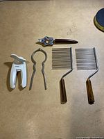 Full view of the six kitchen utensils on a flat surface, showing a white plastic bag clip, metal can opener with brown handle, metal tong, and two metal potato mashers.