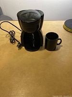 Front view of black single cup coffee maker with power cord and small black ceramic mug to the right.