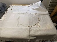 Unfolded white table covering with floral lace cut-out design in center and lace edging, placed on a table