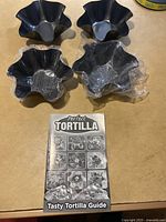 Four black scalloped tortilla baking bowls laid out on a surface with the recipe guide booklet centered below them. Two bowls are unwrapped, two wrapped in plastic.