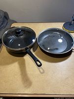 Photo of two frying pans side by side, both with glass lids and black handles, showing used but good condition.