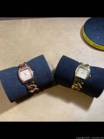 Two watches displayed side-by-side on black foam cushions, one gold-tone and one rose gold-tone, both with curved rectangular faces and chain link bracelets.