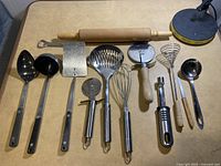 Photo showing various cooking utensils including a wooden rolling pin, metal ladles, whisks, pizza cutters, and spatulas arranged on a surface.