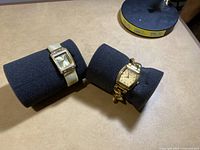 Two wrist watches displayed on black cushions, one with a white band and rectangular case, the other with a gold-tone bracelet and rectangular case.
