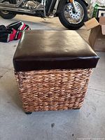 Front angle view of the wicker storage ottoman with the brown cushioned lid.