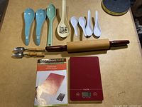 Overview photo showing all items in the lot, including Wolfgang Puck digital kitchen scale, instruction manual, rolling pin, various plastic and bamboo spoons and forks laid on table.